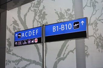 A directional airport sign displaying terminal and gate information against a perforated wall with a tree pattern. The sign indicates terminals ACDEF and gates B1-B10 with icons for boarding and traveler services.