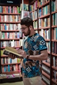 A man with a beard and glasses wearing earphones is engrossed in reading a book. He is standing in a library or bookstore, surrounded by bookshelves filled with a variety of books. The shelves are packed tightly with books of different colors and sizes.
