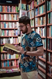 A man with a beard and glasses wearing earphones is engrossed in reading a book. He is standing in a library or bookstore, surrounded by bookshelves filled with a variety of books. The shelves are packed tightly with books of different colors and sizes.