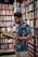 A man with a beard and glasses wearing earphones is engrossed in reading a book. He is standing in a library or bookstore, surrounded by bookshelves filled with a variety of books. The shelves are packed tightly with books of different colors and sizes.