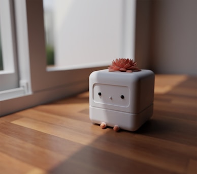 A small, cube-shaped robot with a simple face stands on a wooden surface near a window. It has a flower-like decoration on its top and small, round stubs resembling feet at its base.