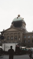 A historic building with a large dome is undergoing renovation, as evidenced by the scaffolding surrounding the dome. The architectural details include ornate columns and statues. Several vehicles, including trucks and bicycles, are on the street in front of the building, with pedestrians walking by.