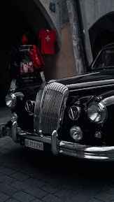An elegant vintage car with a polished black exterior is parked on a cobblestone street. Its prominent chrome grille and classic headlights exude a sense of style and history. Above, a red shirt with a Swiss flag is displayed on a wall, adding a pop of color.