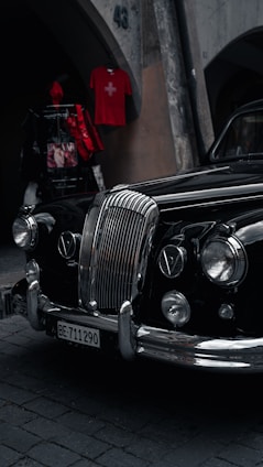 An elegant vintage car with a polished black exterior is parked on a cobblestone street. Its prominent chrome grille and classic headlights exude a sense of style and history. Above, a red shirt with a Swiss flag is displayed on a wall, adding a pop of color.