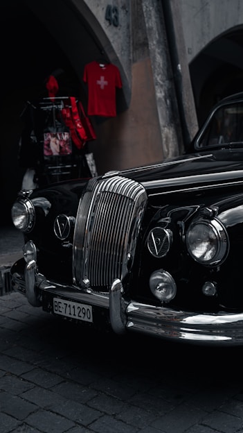 An elegant vintage car with a polished black exterior is parked on a cobblestone street. Its prominent chrome grille and classic headlights exude a sense of style and history. Above, a red shirt with a Swiss flag is displayed on a wall, adding a pop of color.