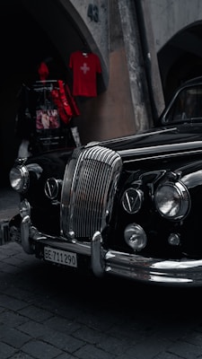 An elegant vintage car with a polished black exterior is parked on a cobblestone street. Its prominent chrome grille and classic headlights exude a sense of style and history. Above, a red shirt with a Swiss flag is displayed on a wall, adding a pop of color.