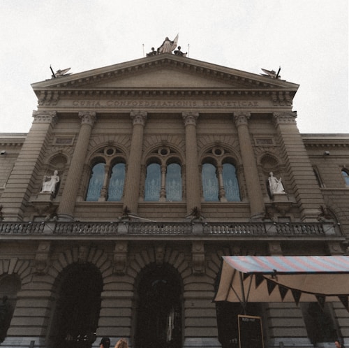 A historic, neoclassical building with ornate columns and statues adorning the facade. The inscription reads 'Curia Confederationis Helveticae'. The architecture features multiple arched windows and decorative elements, suggesting a government or significant institutional structure. There is a tent or awning in the foreground with people gathered beneath.