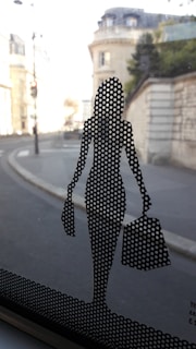 A perforated silhouette of a stylish woman carrying shopping bags is adhered to a glass window. Beyond the glass, a blurred urban street view with historic architecture is visible.