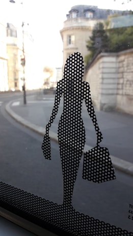 A perforated silhouette of a stylish woman carrying shopping bags is adhered to a glass window. Beyond the glass, a blurred urban street view with historic architecture is visible.