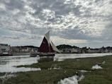 A traditional sailing boat with large sails is navigating a calm river in front of a row of quaint houses. The cloudy sky suggests an overcast day, casting a serene and peaceful atmosphere over the picturesque village setting. Grassy banks line the river, adding a touch of natural beauty to the scene.