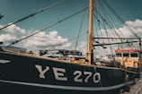 A large fishing boat with the markings 'YE 270' docked at a marina. The vessel has numerous ropes and rigging against a backdrop of a cloudy sky. Other boats and a marina structure are visible in the background.