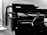A monochrome image of a black multifunction printer with the brand name visible on the top. The printer displays a control panel and an open paper tray. It is positioned against a plain wall with a small portion of a person or object partially visible in the foreground.