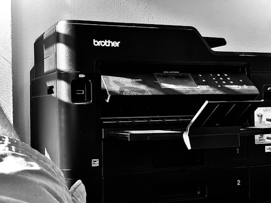 A monochrome image of a black multifunction printer with the brand name visible on the top. The printer displays a control panel and an open paper tray. It is positioned against a plain wall with a small portion of a person or object partially visible in the foreground.