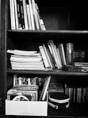 A wooden bookshelf filled with an assortment of books, papers, and DVDs in a box. The books and papers are organized in rows, while the DVDs are neatly arranged in a box at the bottom. The image is in black and white, highlighting the textures of the paper and wood.