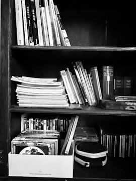 A wooden bookshelf filled with an assortment of books, papers, and DVDs in a box. The books and papers are organized in rows, while the DVDs are neatly arranged in a box at the bottom. The image is in black and white, highlighting the textures of the paper and wood.