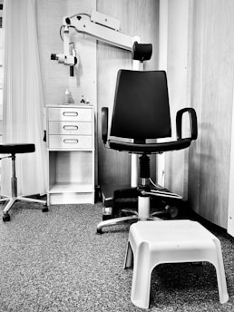 A monochrome image of a clinical or office setting featuring a black swivel chair with armrests and a rolling base. Next to the chair is a small white plastic step stool. In the background, there is shelving with a few items on it, including bottles and a dispenser. A piece of equipment with an adjustable arm is attached to the wall, likely used for medical or professional purposes. The floor is carpeted, and there is a curtain partially visible on the left side.