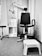 A monochrome image of a clinical or office setting featuring a black swivel chair with armrests and a rolling base. Next to the chair is a small white plastic step stool. In the background, there is shelving with a few items on it, including bottles and a dispenser. A piece of equipment with an adjustable arm is attached to the wall, likely used for medical or professional purposes. The floor is carpeted, and there is a curtain partially visible on the left side.