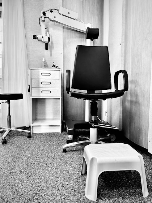 A monochrome image of a clinical or office setting featuring a black swivel chair with armrests and a rolling base. Next to the chair is a small white plastic step stool. In the background, there is shelving with a few items on it, including bottles and a dispenser. A piece of equipment with an adjustable arm is attached to the wall, likely used for medical or professional purposes. The floor is carpeted, and there is a curtain partially visible on the left side.