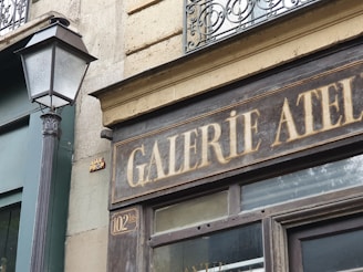 A vintage storefront with a sign reading 'GALERIE ATELIER' in faded white letters. The building has a rustic stone facade with an intricate wrought iron balcony railing above. A classic street lamp post is visible beside the sign, enhancing the old-world charm. The overall appearance suggests a historical or artistic setting.