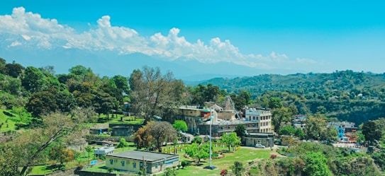 A scenic landscape featuring a small town surrounded by lush green trees and hills. In the background, snow-capped mountains stand majestically under a bright blue sky with fluffy white clouds. The town includes several buildings and a prominent flagpole with the Indian flag visible.