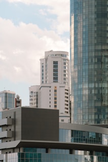 A cluster of modern high-rise buildings with glass facades is set against a backdrop of a partly cloudy sky. The architecture displays a mix of geometric shapes and patterns with reflective surfaces capturing the soft light.