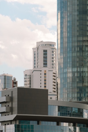 A cluster of modern high-rise buildings with glass facades is set against a backdrop of a partly cloudy sky. The architecture displays a mix of geometric shapes and patterns with reflective surfaces capturing the soft light.
