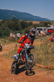 A motocross rider in a red outfit performs a maneuver on a dirt track surrounded by hilly terrain and spectators in the background. The rider is wearing a helmet with blue and red accents, riding a bike with knobby tires suitable for off-road conditions. Some trees and a mountain range are visible under a clear blue sky.