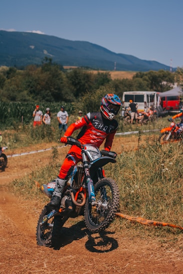 A motocross rider in a red outfit performs a maneuver on a dirt track surrounded by hilly terrain and spectators in the background. The rider is wearing a helmet with blue and red accents, riding a bike with knobby tires suitable for off-road conditions. Some trees and a mountain range are visible under a clear blue sky.