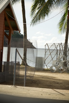 A coastal scene featuring a sandy area with wire mesh fencing and rolls of wire. A small structure with an overhanging roof is visible to the left. Palm trees are situated near the beach, and the ocean can be seen in the background along with a distant hilly landscape.