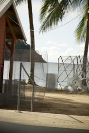 A coastal scene featuring a sandy area with wire mesh fencing and rolls of wire. A small structure with an overhanging roof is visible to the left. Palm trees are situated near the beach, and the ocean can be seen in the background along with a distant hilly landscape.