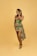 A person stands confidently against a solid yellow background, wearing a vibrant dress with an African print design. The dress features a mix of green and orange colors with intricate patterns. The individual is smiling broadly and appears to be posing with one hand near their face, exuding joy and confidence. They are wearing stylish high-heeled sandals.