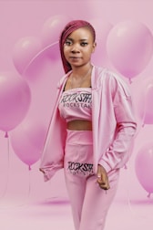A person dressed in a pink outfit with 'Rockstar' printed on the top and pants, standing against a background filled with pink balloons. The individual has braided red hair and is looking directly at the camera.