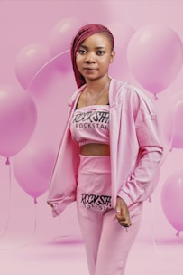 A person dressed in a pink outfit with 'Rockstar' printed on the top and pants, standing against a background filled with pink balloons. The individual has braided red hair and is looking directly at the camera.