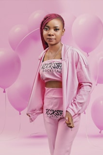 A person dressed in a pink outfit with 'Rockstar' printed on the top and pants, standing against a background filled with pink balloons. The individual has braided red hair and is looking directly at the camera.