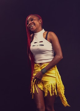 A person with long red braided hair wears a white sleeveless top with cutout details and yellow fringe shorts. The person is smiling and standing against a dark background.