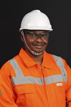 A person wearing an orange safety work uniform with reflective stripes and a white hard hat. The individual is also wearing safety glasses and appears to be smiling.