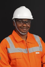 A person wearing an orange safety work uniform with reflective stripes and a white hard hat. The individual is also wearing safety glasses and appears to be smiling.