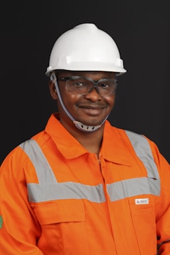 A person wearing an orange safety work uniform with reflective stripes and a white hard hat. The individual is also wearing safety glasses and appears to be smiling.