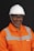 A person wearing an orange safety work uniform with reflective stripes and a white hard hat. The individual is also wearing safety glasses and appears to be smiling.