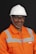 A person wearing an orange safety work uniform with reflective stripes and a white hard hat. The individual is also wearing safety glasses and appears to be smiling.