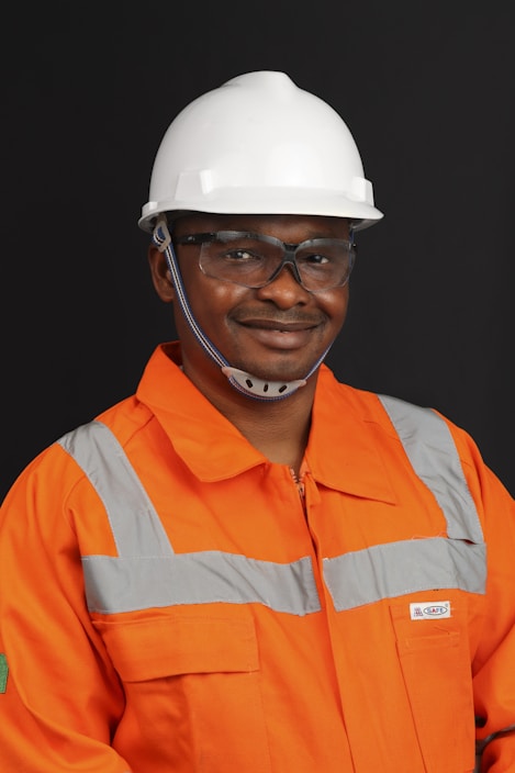 A person wearing an orange safety work uniform with reflective stripes and a white hard hat. The individual is also wearing safety glasses and appears to be smiling.