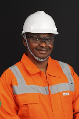 A person wearing an orange safety work uniform with reflective stripes and a white hard hat. The individual is also wearing safety glasses and appears to be smiling.