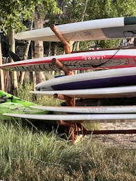 A stack of surfboards and paddleboards is arranged on a wooden rack. The boards have various colors and designs, including red and white, and are placed in an outdoor setting surrounded by grass and trees. Sunlight filters through the leaves, casting shadows.