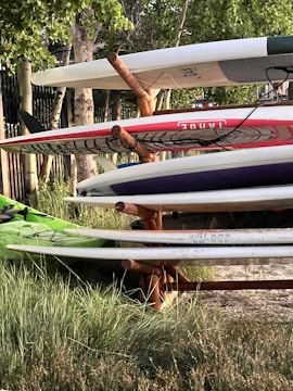 A stack of surfboards and paddleboards is arranged on a wooden rack. The boards have various colors and designs, including red and white, and are placed in an outdoor setting surrounded by grass and trees. Sunlight filters through the leaves, casting shadows.