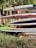 A stack of surfboards and paddleboards is arranged on a wooden rack. The boards have various colors and designs, including red and white, and are placed in an outdoor setting surrounded by grass and trees. Sunlight filters through the leaves, casting shadows.