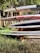 A stack of surfboards and paddleboards is arranged on a wooden rack. The boards have various colors and designs, including red and white, and are placed in an outdoor setting surrounded by grass and trees. Sunlight filters through the leaves, casting shadows.
