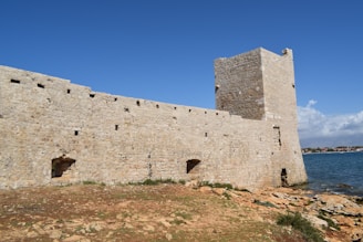 A stone fortress wall stretches across the foreground, with a tall, square tower rising prominently on the right. The fortress overlooks a body of water, likely the sea, under a clear blue sky. Sparse vegetation grows near the base of the wall on rocky terrain.