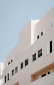 A modern building with minimalist architectural design, featuring beige walls and several rectangular windows with some open shutters. The background is a clear blue sky.