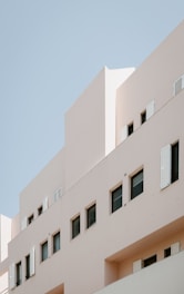 A modern building with minimalist architectural design, featuring beige walls and several rectangular windows with some open shutters. The background is a clear blue sky.