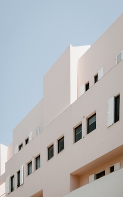 A modern building with minimalist architectural design, featuring beige walls and several rectangular windows with some open shutters. The background is a clear blue sky.